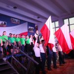 Na scenie uczniowie z klas I – III i ósmoklasiści z podniesionymi flagami Polski