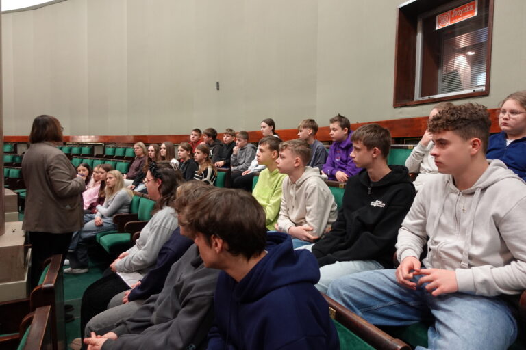 Uczestnicy wycieczki słuchają opowieści przewodnika o pracy posła na galerii Sali Sejmu RP.